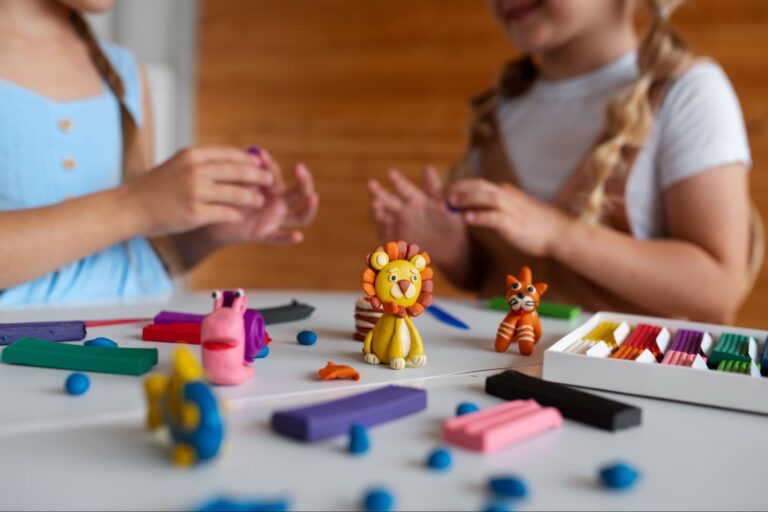 Duas crianças brincando de massinha em frente a uma superfície branca. Existem alguns animais feitos de massinha com alguns pedaços coloridos espalhados.