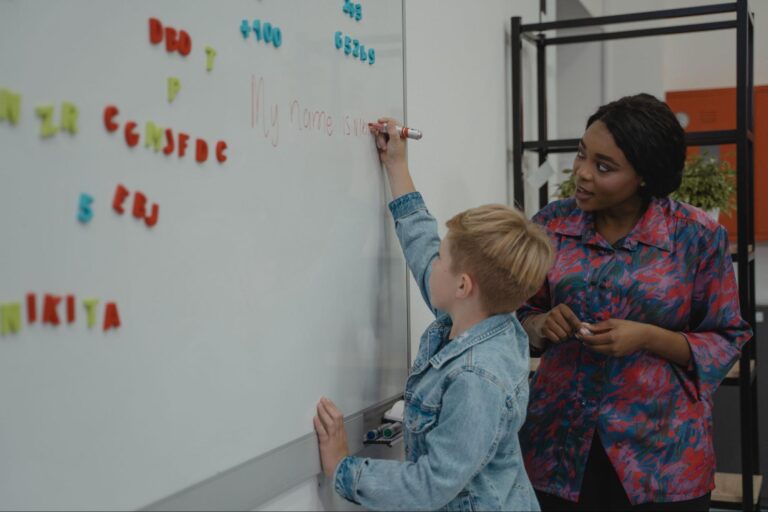 Na imagem, uma professora observa de perto um aluno que escreve na lousa como parte de um projeto de intervenção pedagógica.