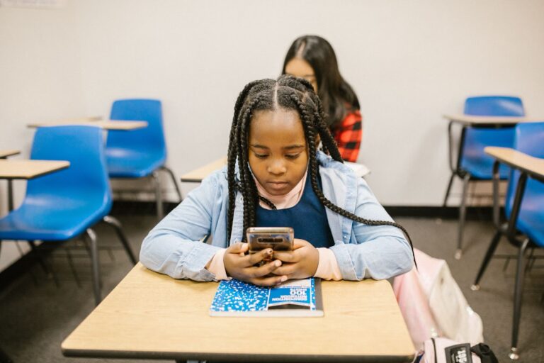 Garota negra de tranças utiliza um smartphone enquanto está sentada em uma carteira na sala de aula.