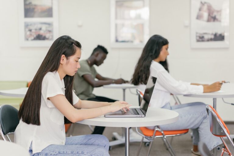 Sala de aula com três estudantes; em primeiro plano vemos uma mulher de cabelos longos usando um notebook e atrás há um homem e uma mulher escrevendo em seus respectivos cadernos. 