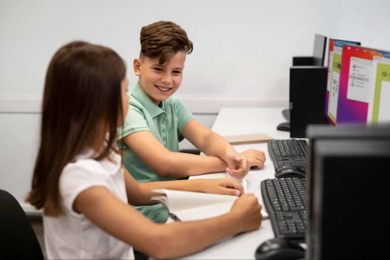 Duas crianças estão conversando sentadas uma ao lado da outra, a sua frente existem alguns computadores indicando que estão no laboratório de informática.