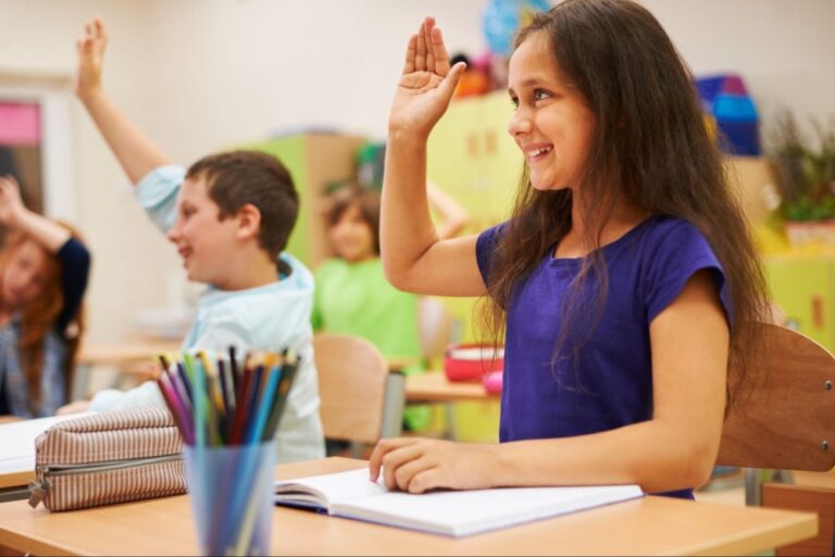 Crianças com uma das mãos levantadas em sala de aula. Há uma criança de azul sorrindo em foco.