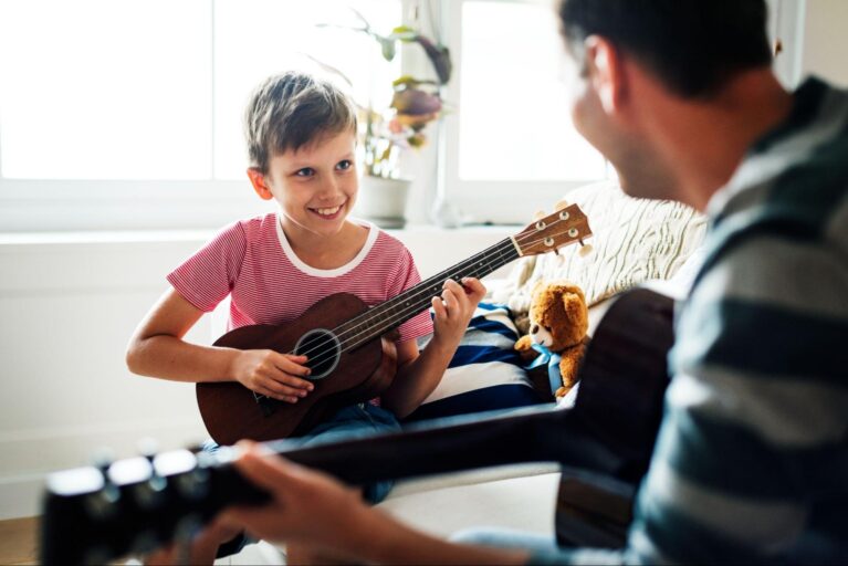 O menino está segurando um violão enquanto olha um adulto à sua frente, também com um instrumento. A imagem representa a inteligência musical.