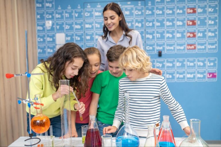 Quatro crianças estão reunidas em frente a uma mesa com materiais da aula de química. A professora está atrás deles observando o procedimento enquanto sorri. 