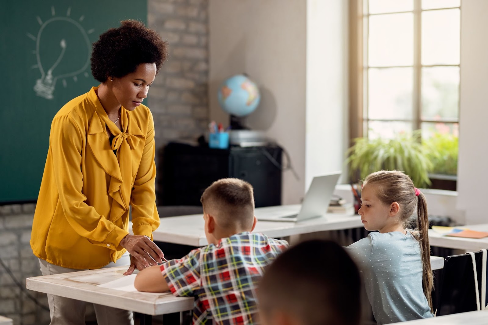A professora está em frente a uma mesa com dois alunos, ela está segurando uma caneta próxima à folha do menino. A imagem representa como trabalhar a autoavaliação em sala de aula.