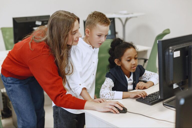 Professora está ao lado de dois alunos, estão em uma sala de informática usando um computador. 