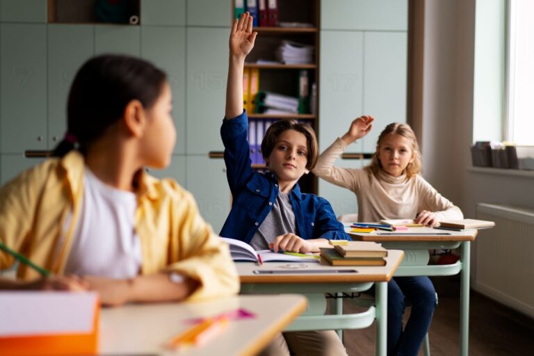 Os alunos estão sentados em mesas separadas, um menino e uma menina estão levantando a mão para tirar suas dúvidas com o professor. A imagem representa o rendimento escolar.