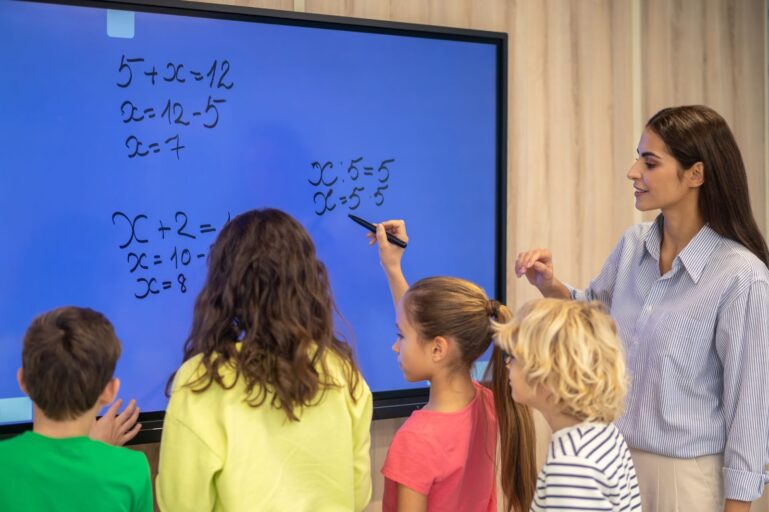 A professora e os alunos estão em frente a uma tela, uma das alunas segura uma caneta para completar a conta de matemática.