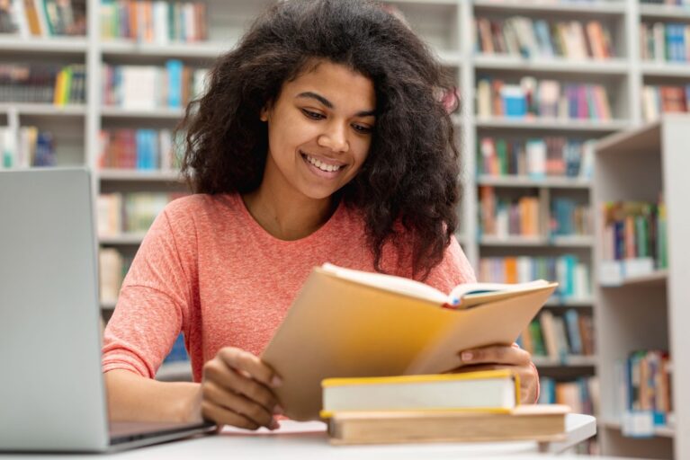 Uma menina está sentada em uma biblioteca, segura um livro em suas mãos enquanto deixa outros dois na mesa do lado oposto que está um notebook.