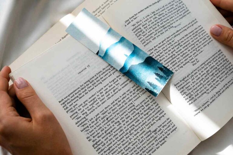 Imagem mostra uma pessoa lendo um livro, segurando-o com as duas mãos. Há um marcador de texto azul posicionado entre as duas páginas abertas.