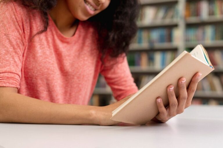 Imagem mostra uma mulher, em um ambiente de uma biblioteca, lendo um livro, apoiada em uma mesa. Ela está com uma expressão sorridente.