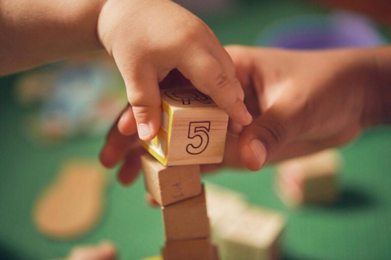 A imagem mostra uma criança e uma pessoa adulta empilhando blocos, um deles bem no centro do enquadramento, mostrando o número “5”.