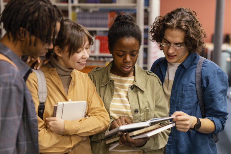 4 estudantes estudando juntos na biblioteca com roupas de tom amarelo, verde e azul.