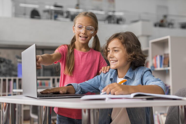 crianças estudando no computador. menino branco com jaqueta jeans, blusa branca, e cabelo ondulado claro, sentado na cadeira com sorriso olhando o notebook. Garota branca de blusa rosa, de cabelo loiro comprido, com oculos redondo apontando para notebook.